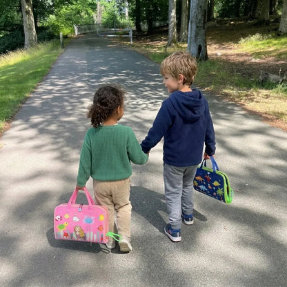 Due bambini camminano mano nella mano in un parco portando le valigette Manine rosa e blu